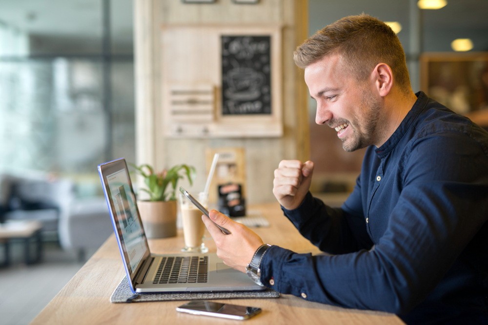 Man with a laptop in front of him celebrating when checking his phone at a coffe shop